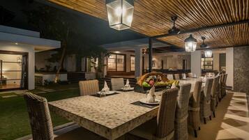 An outdoor dining area with a long table and wicker chairs, under a wooden ceiling with hanging lanterns.