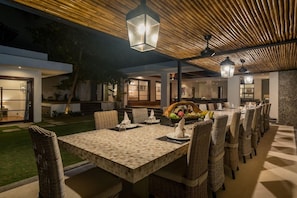 Uma área de refeições ao ar livre com uma mesa comprida e cadeiras de vime, teto de madeira e luminária estilo lanterna.