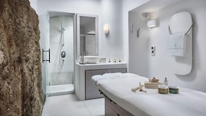 A modern bathroom with a glass-enclosed shower, a vanity with a mirror, and a massage table with toiletries. - Belvedere Waterfront Villa and Suites (Mykonos)