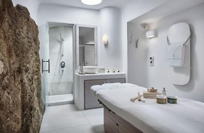 Ein modernes Badezimmer mit einer einteilig verglasten Dusche, einem Waschtisch mit Spiegel und einer Massageliege mit Toilettenartikeln.