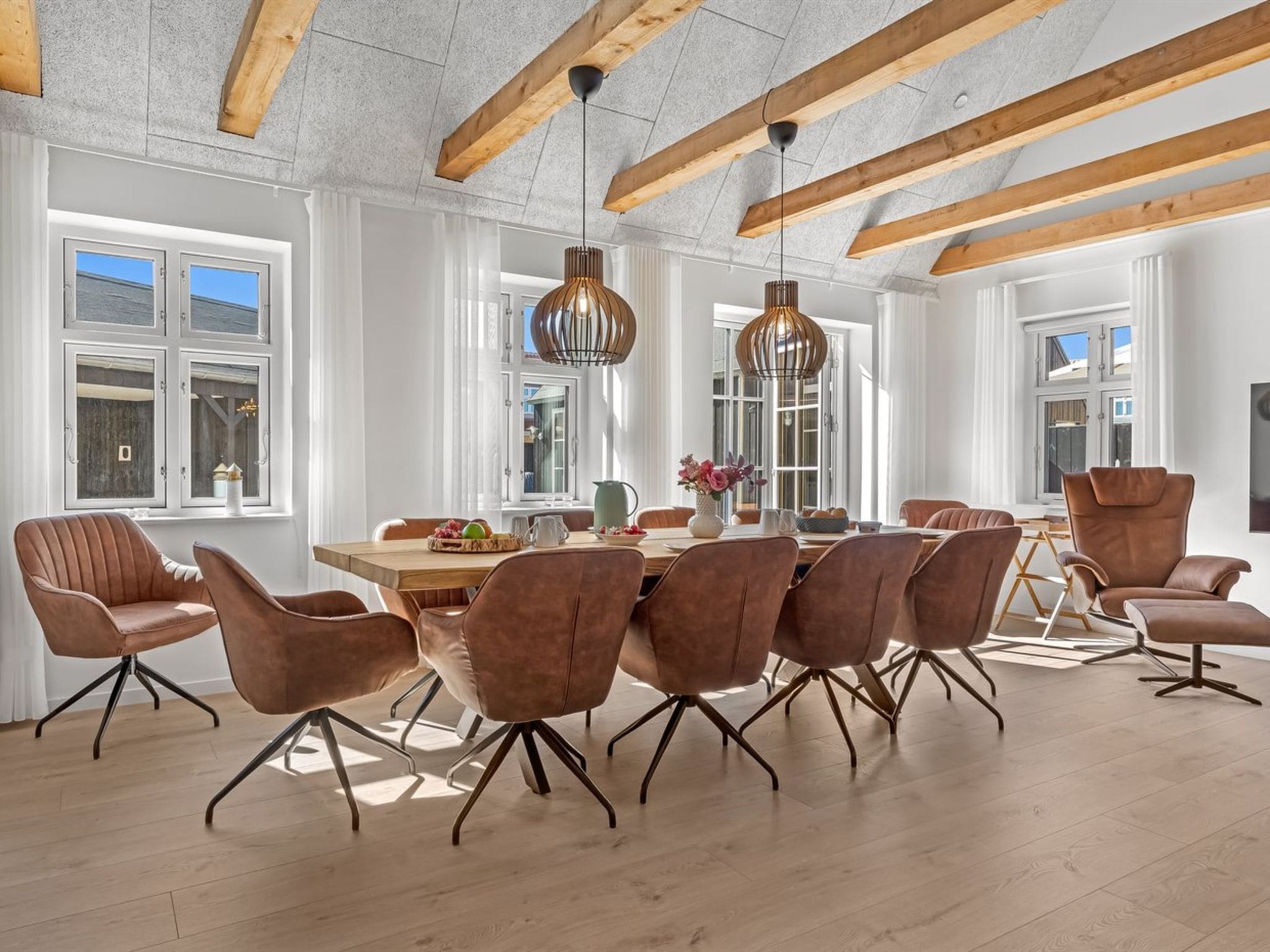 Een ruime eethoek met een houten tafel, moderne stoelen, grote ramen en zichtbare houten balken aan het plafond.