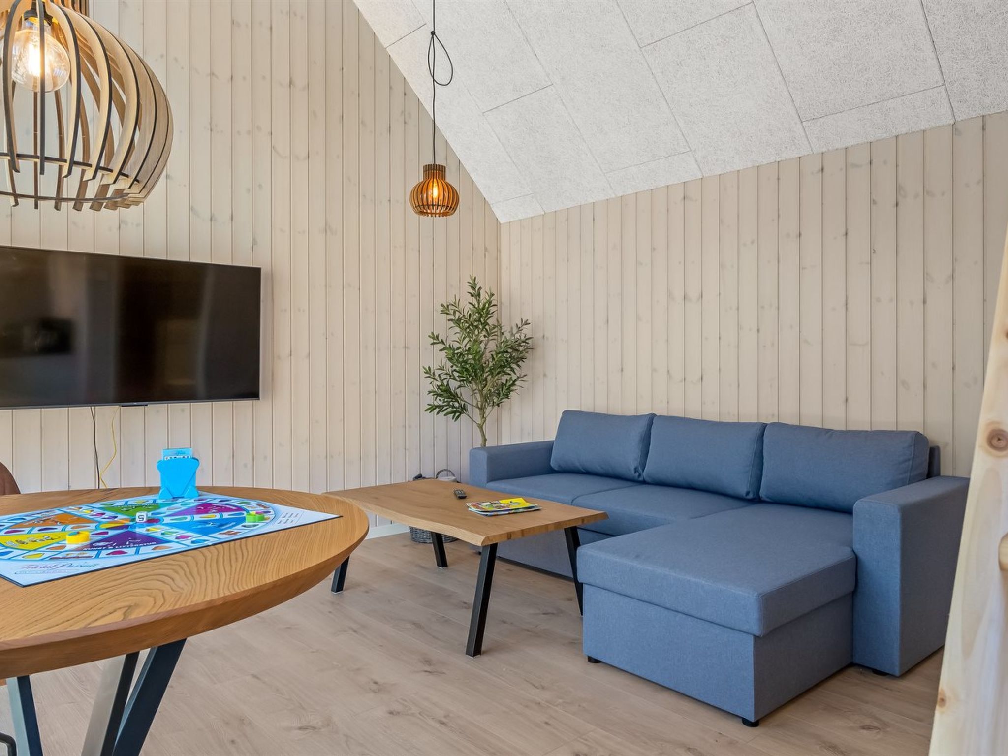 Un salon moderne avec un canapé d’angle, une table basse en bois, un téléviseur à écran plat fixé au mur et un luminaire suspendu.
