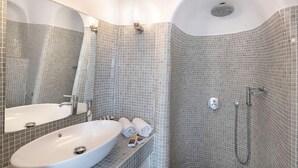 A bathroom with a white sink, a mirror, a shower with a handheld showerhead, and a tiled wall. - Veranda View (Santorini)
