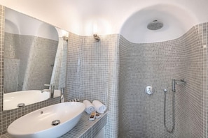 A bathroom with a white sink, a mirror, a shower with a handheld showerhead, and a tiled wall. - Veranda View (Santorini)