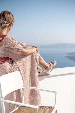 A person sitting on a white chair with a view of a body of water and distant land. - Veranda View (Santorini)