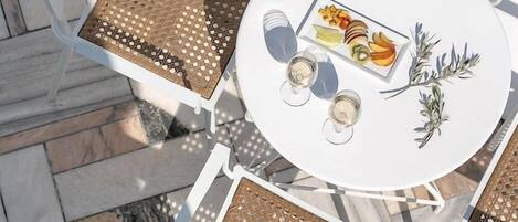 A round table with a white tablecloth, surrounded by white chairs with woven seat cushions, and a plate of fruit and a glass of white wine on the table.