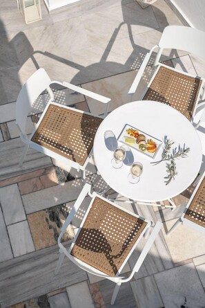 A round table with a white tablecloth, surrounded by white chairs with woven seat cushions, and a plate of fruit and a glass of white wine on the table. - Veranda View (Santorini)