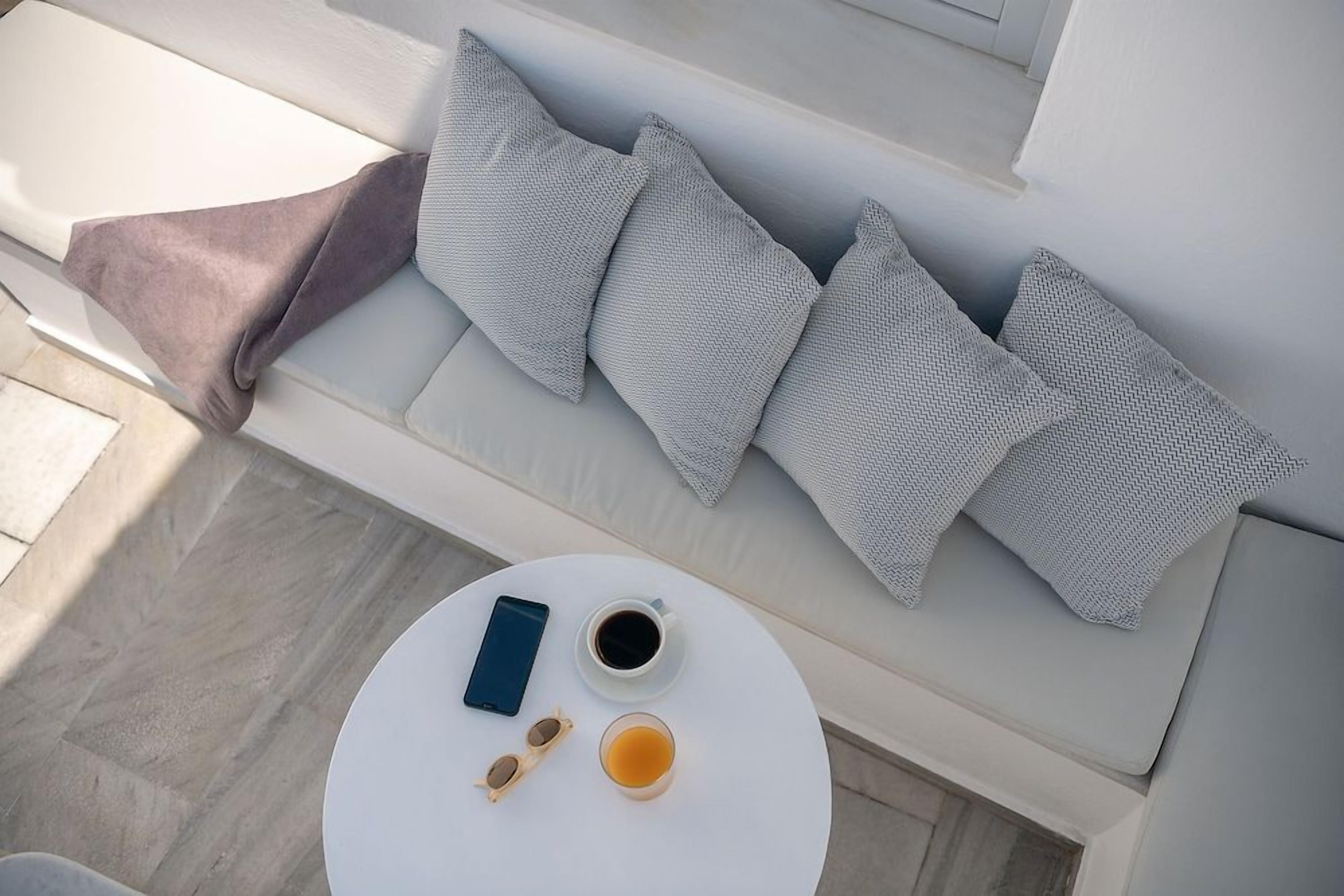 Een witte bank met grijs gestreepte kussens, een ronde witte tafel met een smartphone, een kop koffie en een theeglas.