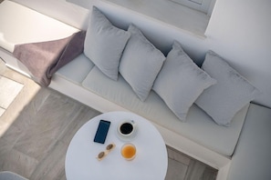 A white sofa with grey patterned cushions, a round white table with a smartphone, a cup of coffee, and a glass of tea. - Veranda View (Santorini)