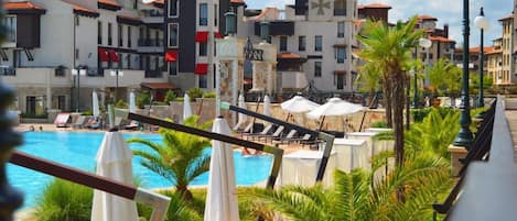 A poolside area with lounge chairs and umbrellas, surrounded by buildings with balconies and red awnings.