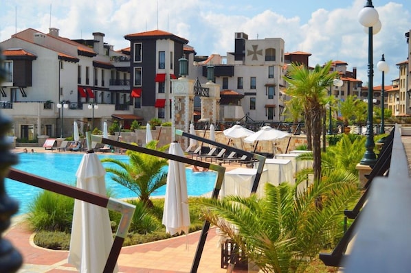 A poolside area with lounge chairs and umbrellas, surrounded by buildings with balconies and red awnings. - Oasis Resort & Spa, Lozenets (Tsarevo)
