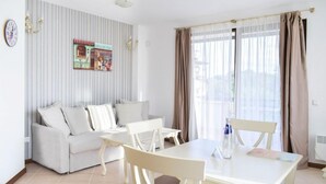 Sala de estar moderna com sofá, mesa de jantar e cadeiras. Há uma janela com cortinas, um relógio de parede e um objeto decorativo sobre o sofá.