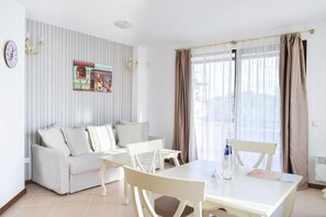 A modern living room with a sofa, dining table, and chairs. There is a window with curtains, a wall clock, and a decorative piece above the sofa. - Oasis Resort & Spa, Lozenets (Tsarevo)