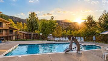 A swimming pool with a lounge chair and a glass of water on the side, surrounded by trees and a mountain in the background.