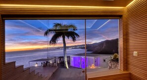 Un interior moderno con un amplio ventanal que da a una puesta de sol costera, una palmera y una escalera que conduce a un espacio exterior.