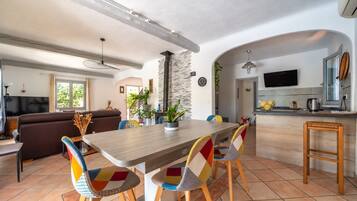 Een moderne keuken met een centraal eiland, een eethoek met kleurrijke stoelen, en een woonkamer met een bank en televisie.