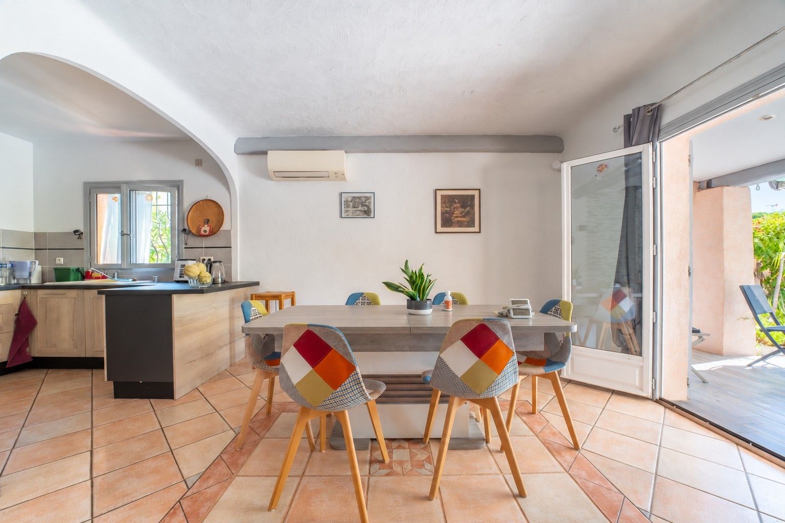 A modern kitchen with a dining area, featuring a large island, chairs with colorful cushions, and a sliding glass door leading to an outdoor area.
