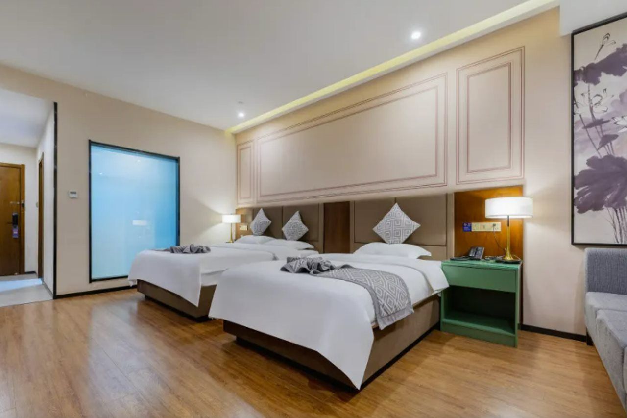 Habitación de hotel con dos camas, una mesita de noche verde, una lámpara de pared y un ventanal grande.