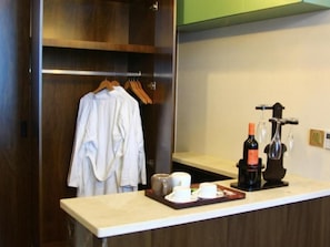 A hotel room with a wooden wardrobe, a minibar with a bottle and glasses, and a counter with a tray of items.