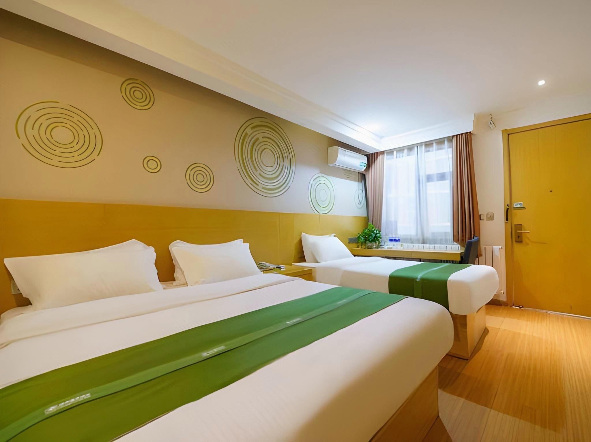 A hotel room with two beds, a window with curtains, a wall-mounted air conditioner, and a wooden door.