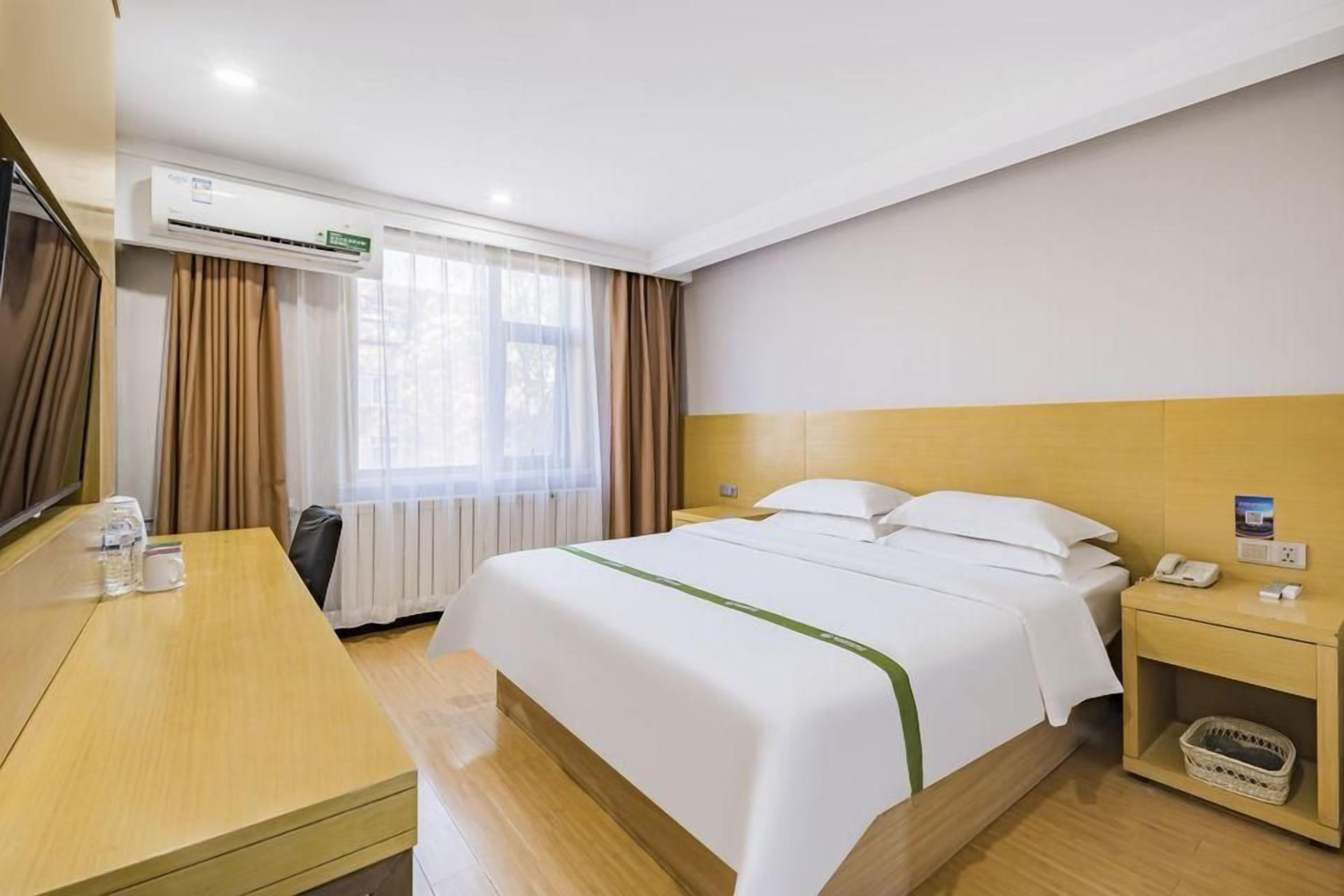A hotel room with a bed, a desk, a chair, a nightstand, a telephone, and an air conditioning unit.