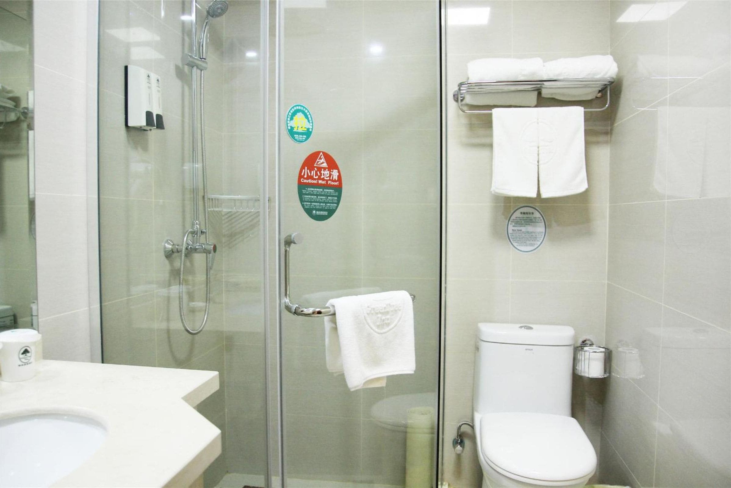 A bathroom with a glass shower enclosure, a toilet, and a sink. There are towels hanging on a rack and a soap dispenser on the wall.