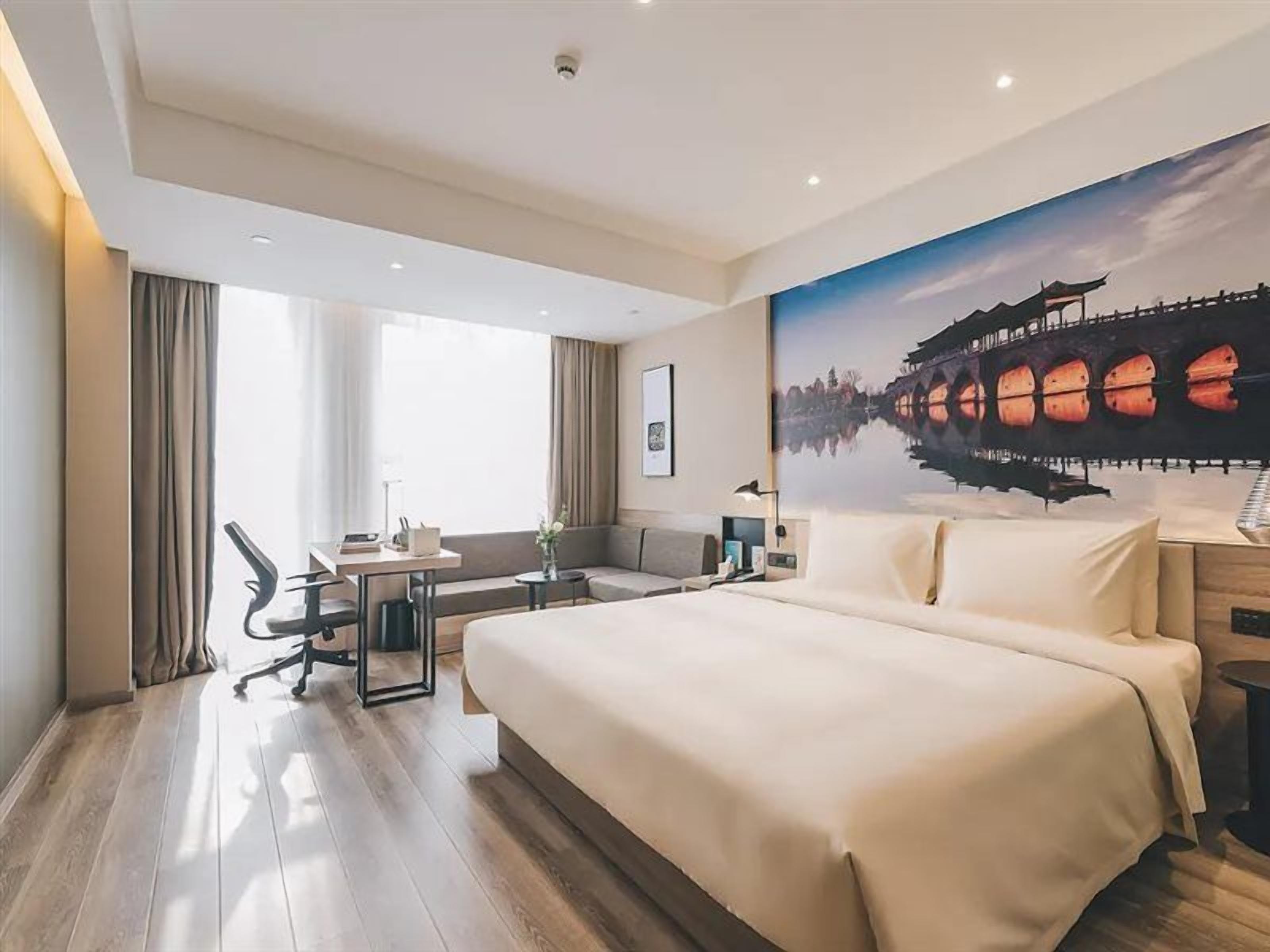 A hotel room with a large bed, a desk, a chair, a sofa, and a painting of a bridge.