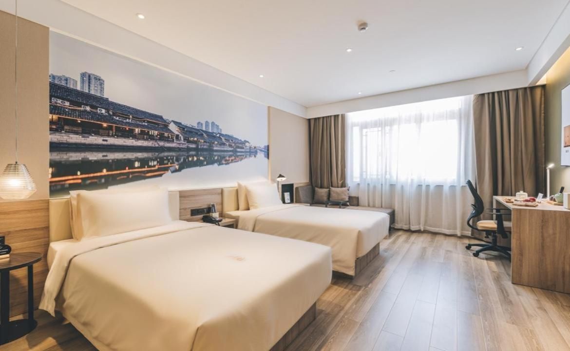 Habitació d'hotel amb dos llits, un escriptori, una cadira, un finestra gran amb cortines i un mural de paisatge urbà a la paret.