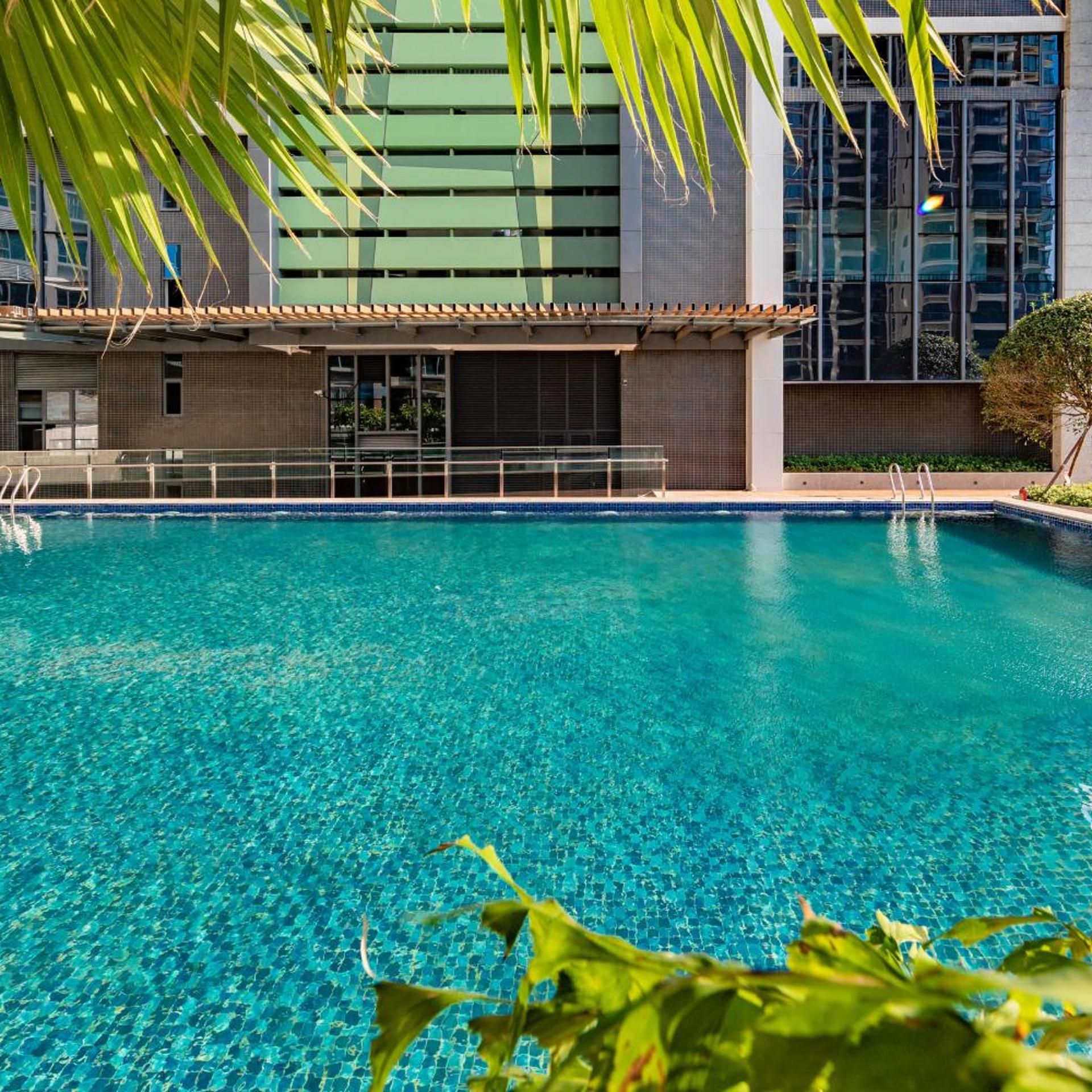 Piscina azul cristalina con borde de azulejos, rodeada por un edificio con amplias ventanas y fachada verde.
