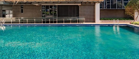 A clear blue pool with a tiled edge, surrounded by a building with large windows and a green facade.