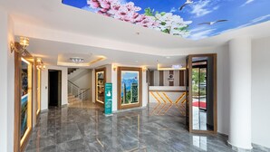 A hotel lobby with a marble floor, a reception desk, a staircase, and a wall with a floral mural. - Theluna Suite Hotel (Konyaalti)