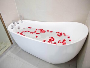 Uma banheira independente branca com pétalas de rosa flutuando na água, uma torneira cromada e piso ladrilhado.