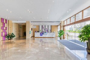Uma área de recepção de hotel com piso de mármore, balcão de atendimento e plantas em vasos.