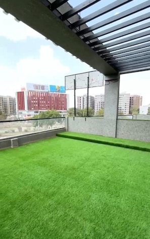 A rooftop terrace with artificial grass, a concrete structure, and a view of surrounding buildings.