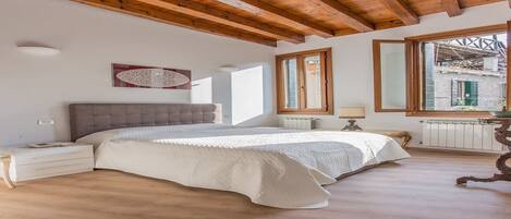 A bedroom with a large bed, wooden ceiling beams, a radiator, and a window with a view of a building outside.