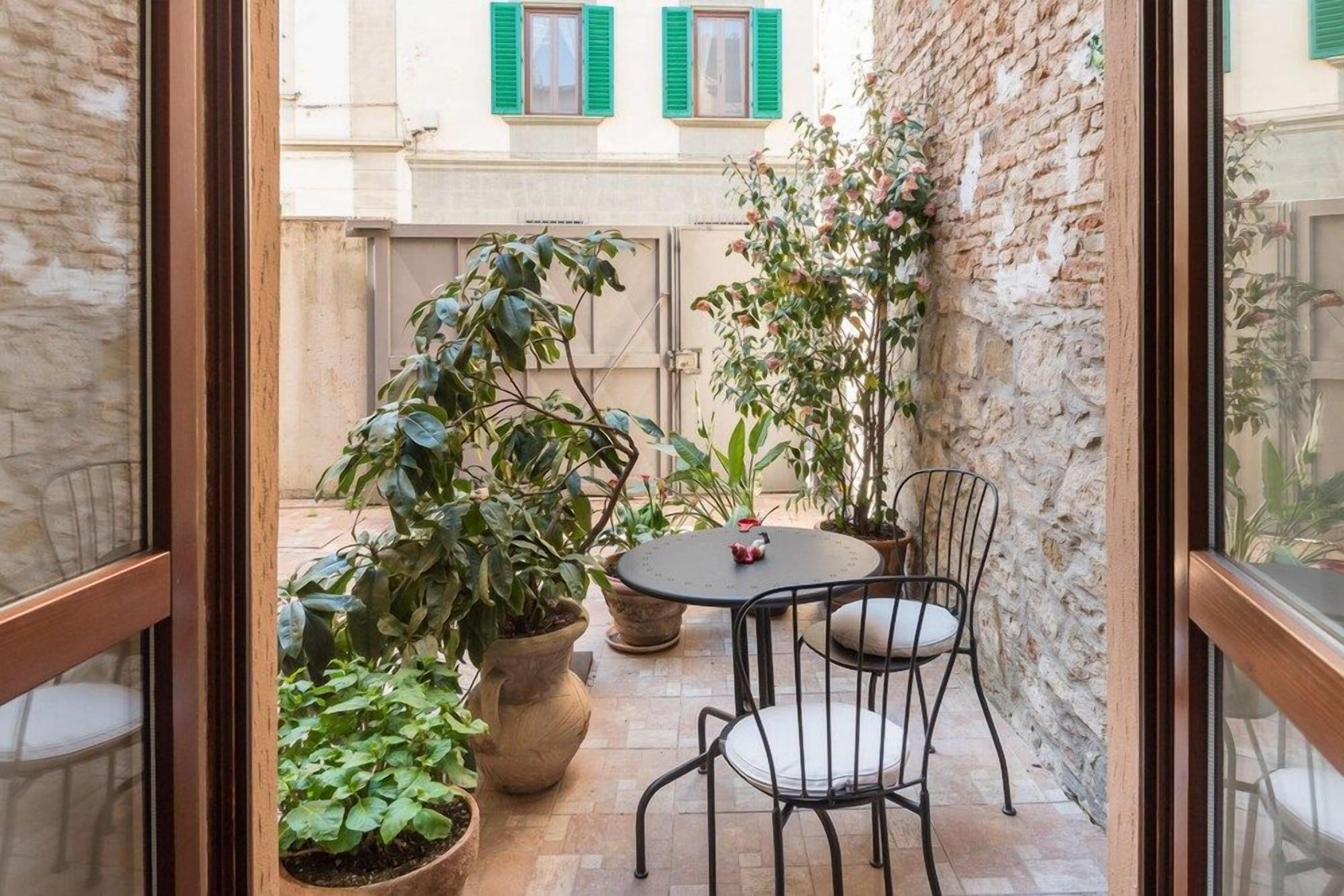 A small outdoor patio with a round table and two chairs, surrounded by potted plants and a brick wall.