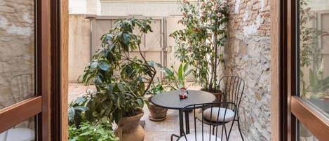 Un pequeño patio al aire libre con una mesa redonda y dos sillas, rodeado de plantas en macetas y un muro de ladrillo.