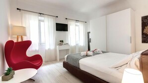 A hotel room with a bed, a red chair, a small table, a TV, and a window with curtains. - Mamo Florence - Velluti Apartment (Florence)