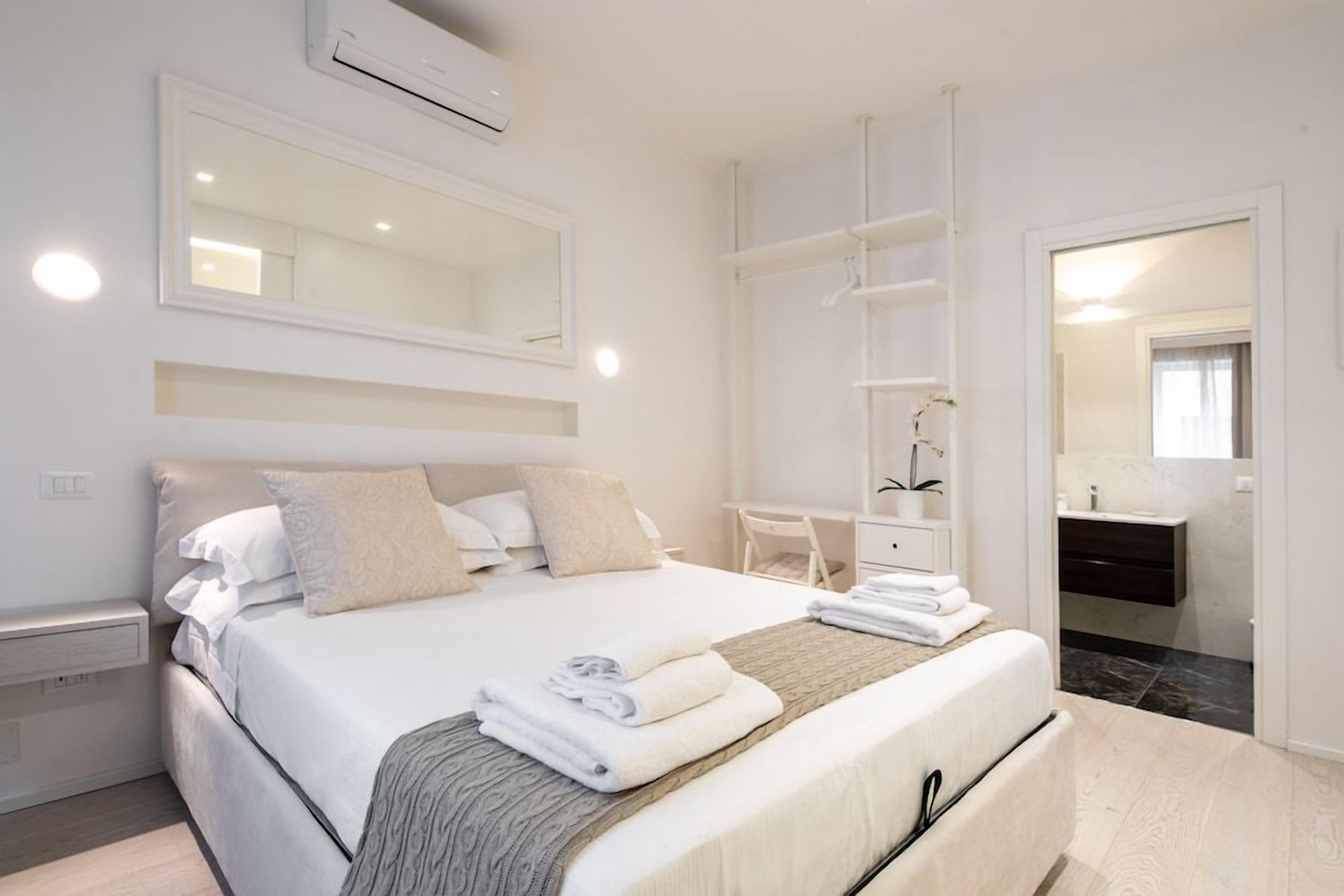 A modern bedroom with a large bed, bedside tables, a wardrobe, and a bathroom visible through an open door.
