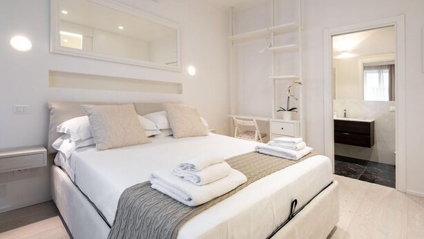 A modern bedroom with a large bed, a bedside desk, a built-in wardrobe, and a bathroom visible through an open door. - Suite Jasmine (Florence)