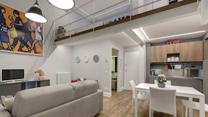 A modern apartment with a grey sofa, dining table, and white chairs. A kitchen with wooden cabinets and a 'Home' sign is visible. - Mamo Florence - St Elizabeth Loft Apartment (Florence)