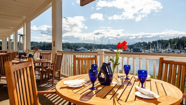 An outdoor dining area with wooden tables and chairs, a vase with a red flower, and a view of boats and trees in the background. - Resort At Port Ludlow (Port Ludlow)