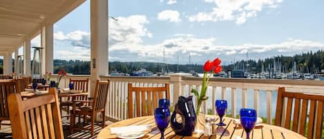 Un área de comedor al aire libre techada con mesas y sillas, techo de madera y vista a barcos y árboles.