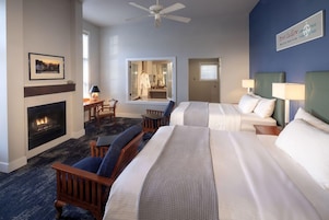A hotel room with a fireplace, two beds, a sitting area with a chair and a table, and a ceiling fan.