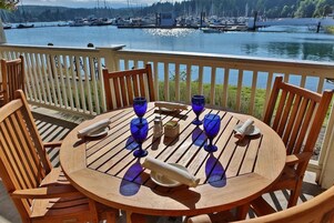 A wooden deck with a round table and chairs, overlooking a marina with boats.