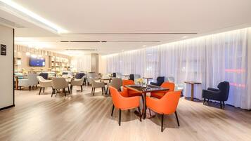 A modern hotel lobby with a dining area featuring orange chairs, a TV, and a bar.
