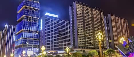 A nighttime cityscape with illuminated high-rise buildings, a water feature with fountains, and a well-lit public square.