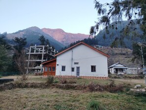 A single-story building with a white facade and a red roof, surrounded by a grassy area with trees and mountains in the background.