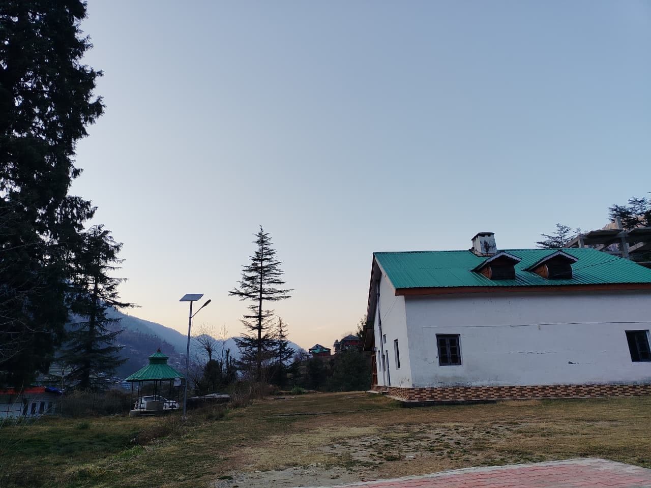 Un edificio blanco con techo verde, rodeado de árboles y una montaña al fondo.