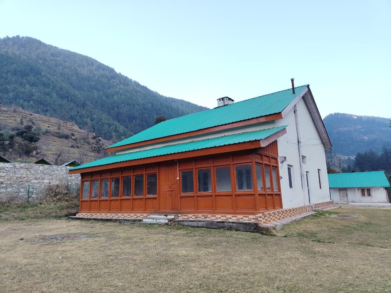 A two-story building with a green roof, wooden exterior, and a stone wall in the background.